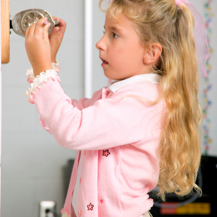 A young girl with long blonde hair, wearing a pink sweater, using a pencil sharpener on the wall. The background features a light-colored wall and some colorful decorations, emphasizing the usability of the sharpener for children